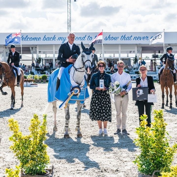 Photo d'un cheval gris avec une couverture bleue et trois personnes a pied avec des trophées dans les mains